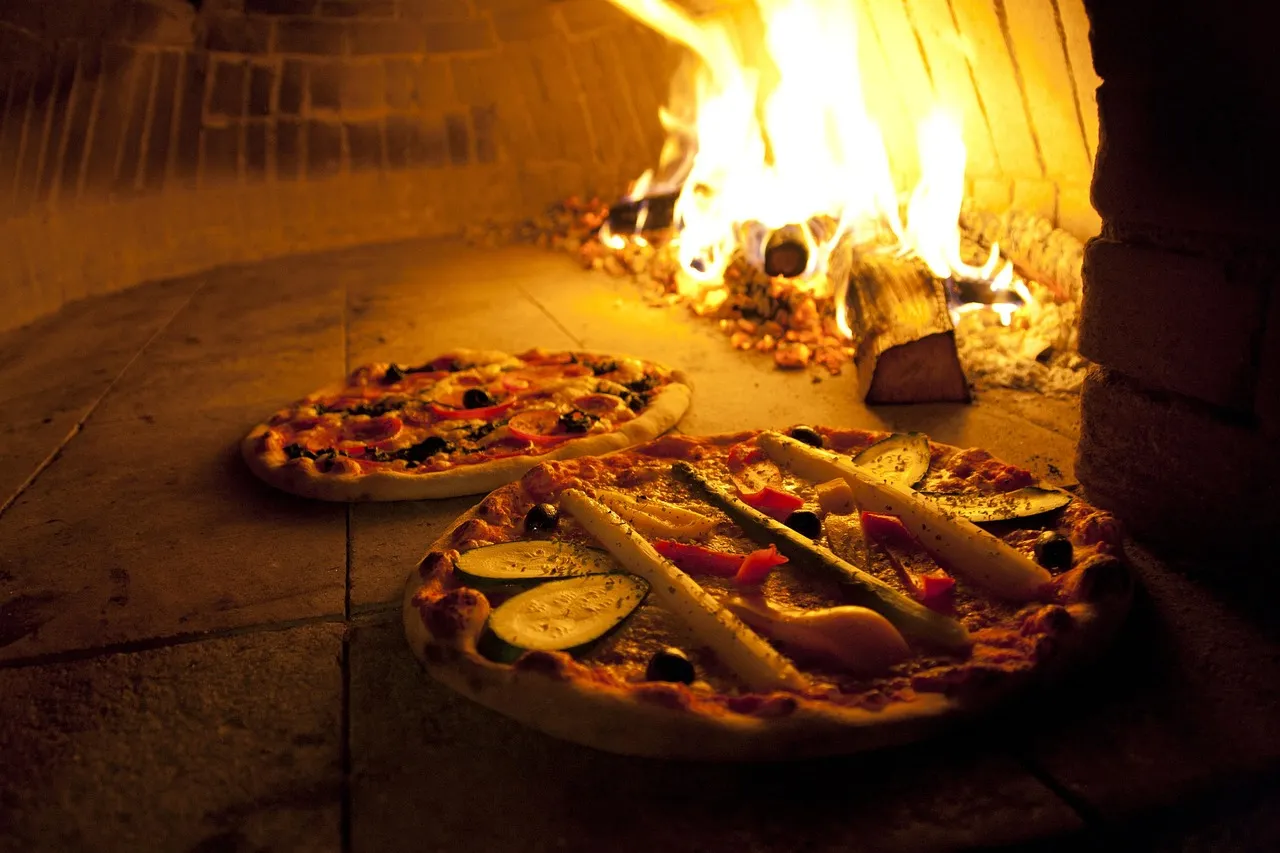 Two pizzas on the stone floor of a brick wood-fired oven, with logs burning brightly at the back