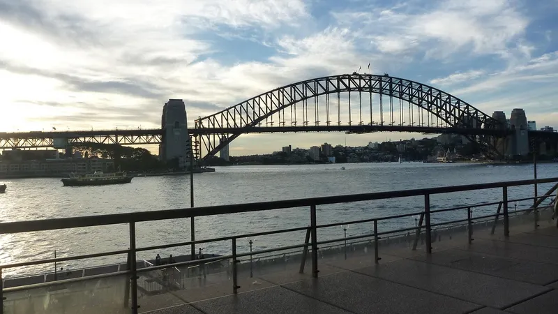 The Sydney Harbour Bridge in late afternoon light, seen across the water from behind a harbourside railing, with a ferry crossing.