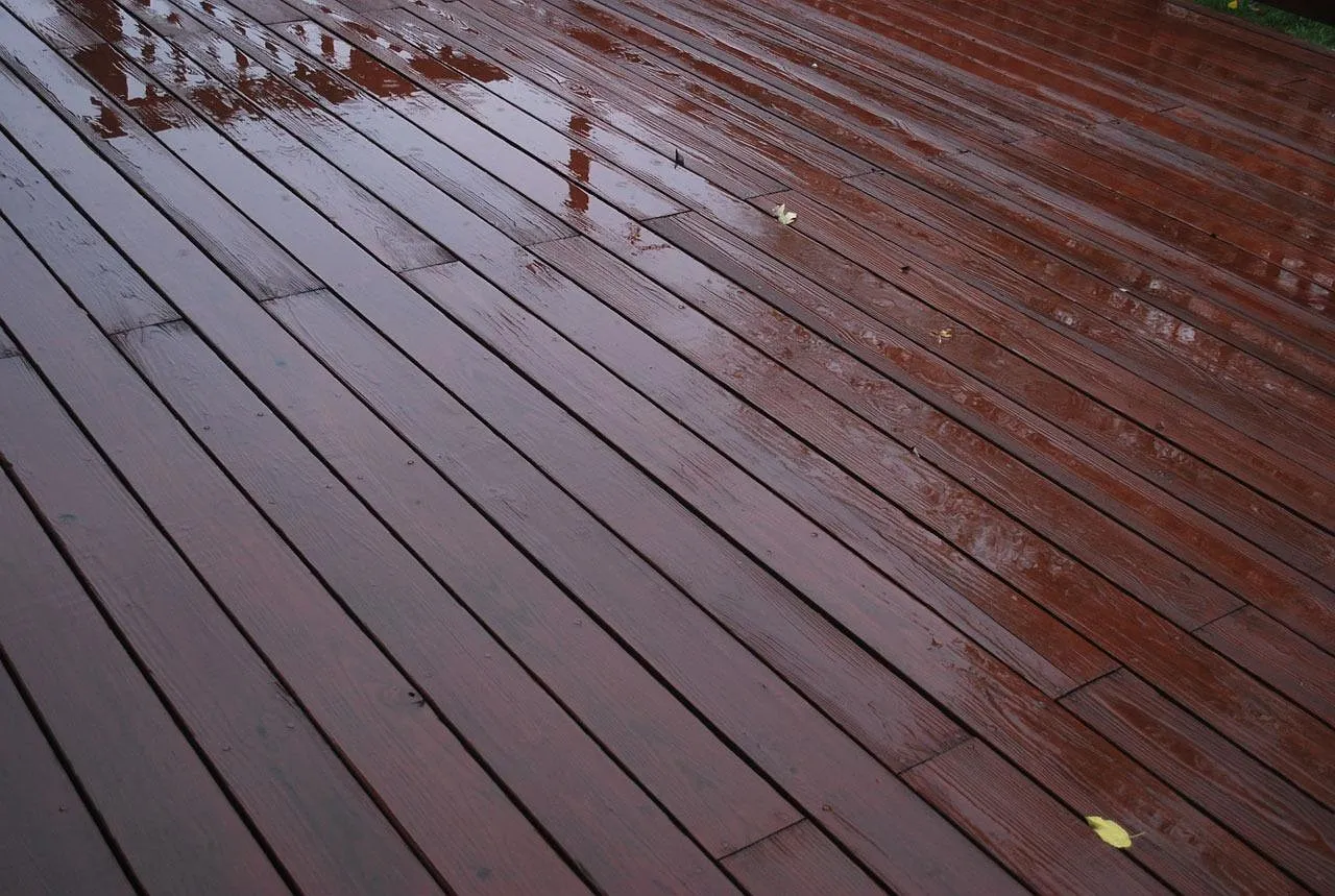 Wet, dark-stained deck boards after rain, with water pooled along the joins and a fallen leaf.
