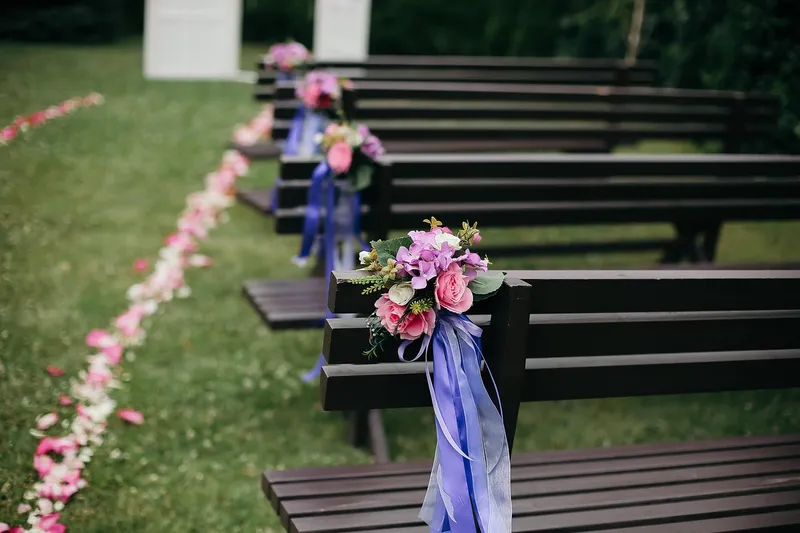 Rows of dark wooden benches on a lawn, each tied with a posy of pink flowers and lilac ribbon, beside an aisle edged with rose petals