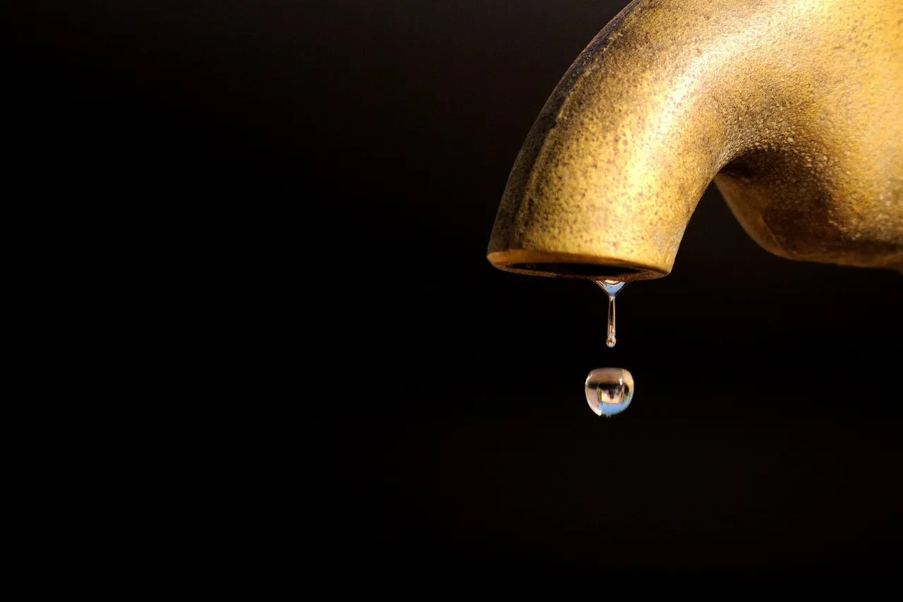 A single drop of water falling from the spout of a brass tap, against a black background.