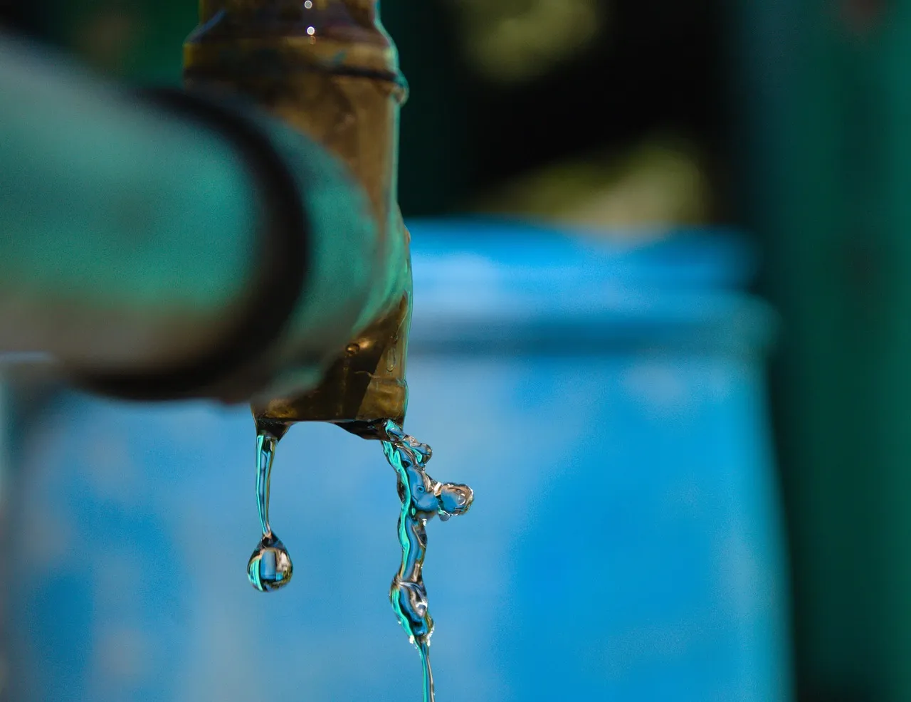 Water dripping and trickling from the end of an outdoor pipe, with a blurred blue drum behind it.