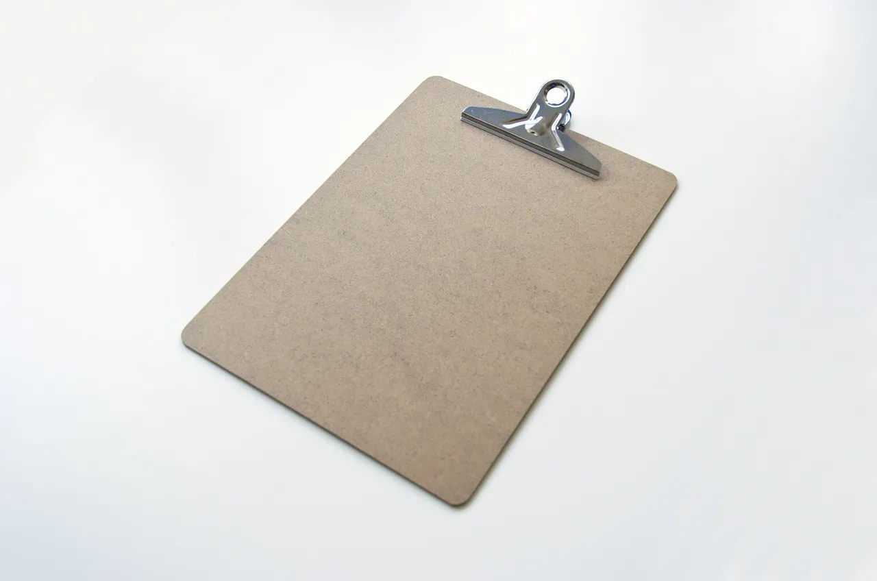 A bare brown hardboard clipboard with a silver metal clip at the top, lying at an angle on a plain white surface. No paper is clipped to it.
