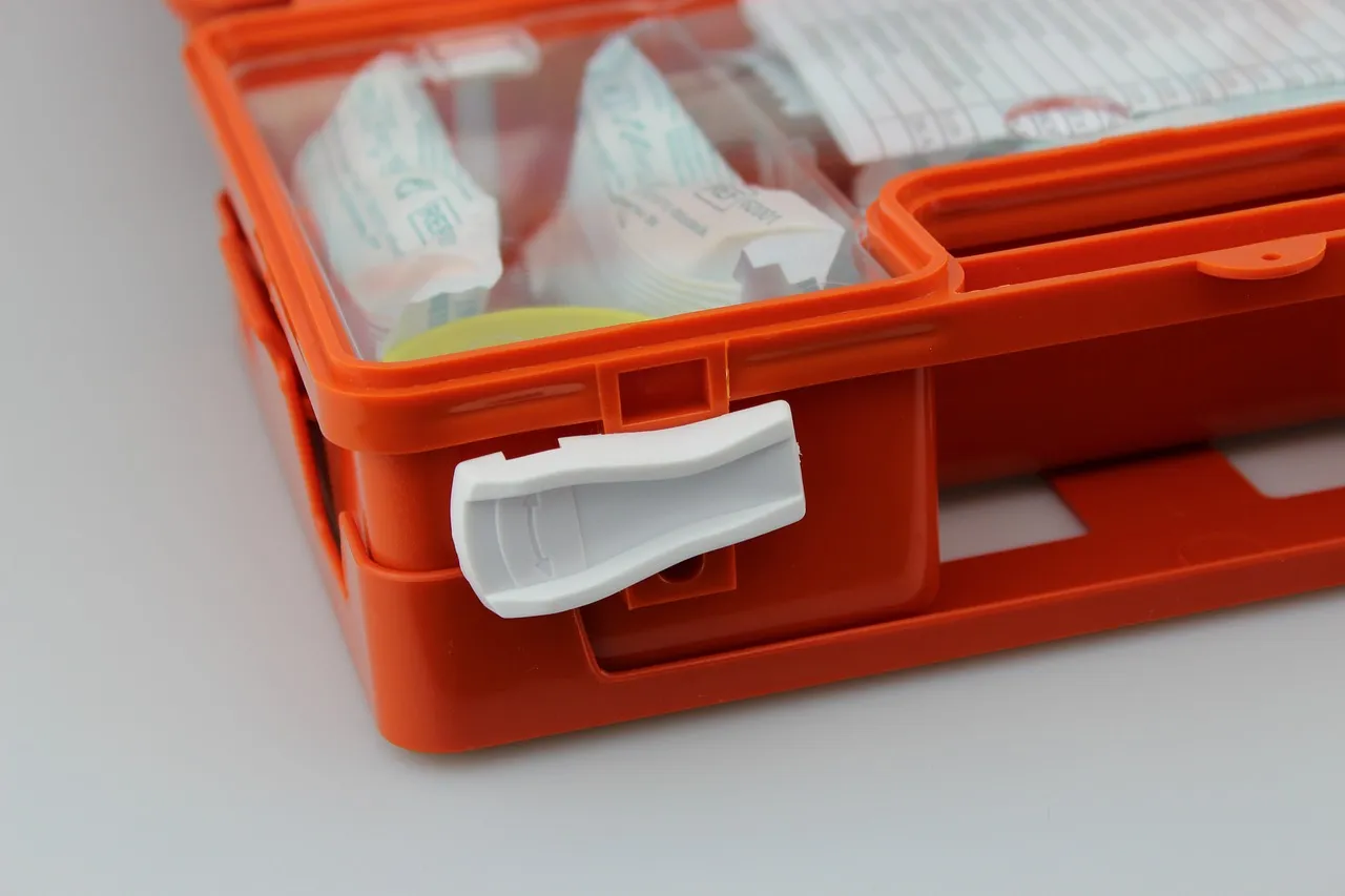 Close view of the corner of an orange plastic first aid case on a pale grey surface. A white catch holds the lid shut, and wrapped dressings and supplies show through the clear lid.